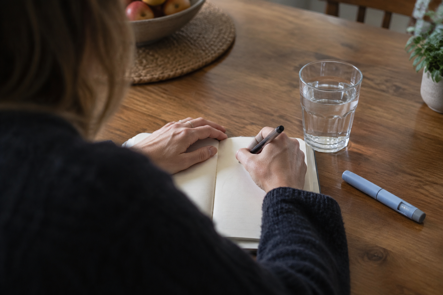 Mounjaro user recording symptoms at a kitchen table beside a glass of water and an unbranded injection pen